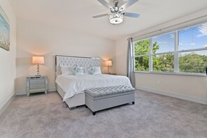 A bedroom with a large bed, two bedside tables, a ceiling fan, and a window with a view of greenery.