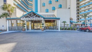 La entrada del hotel Ocean Reef, con un paseo techado, palmeras y un coche rojo aparcado cerca.