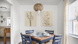 Uma sala de jantar com mesa de madeira posta para quatro pessoas, cercada por cadeiras azuis com assentos trançados. Há duas obras de arte fixadas na parede, retratando cactos e designs abstratos.