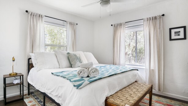 Um quarto com uma cama, mesa de cabeceira e um banquinho.