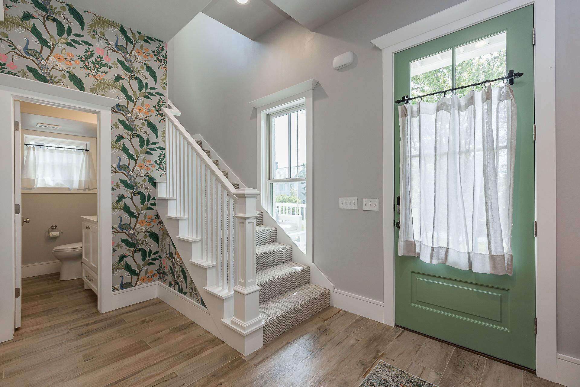 A staircase with white railings and patterned wallpaper, leading to a room with a green door and a window with sheer curtains.