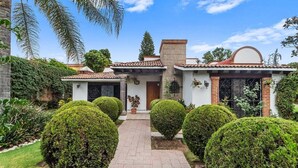 A two-story house with a tiled roof, a brick chimney, and a small porch with potted plants. - +MS +Casa Tequis +Hermoso Jardín (Tequisquiapan)