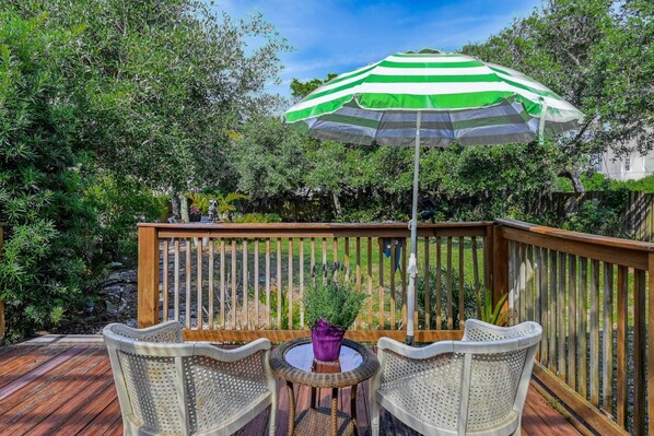 A wooden deck with two wicker chairs and a small round table, a green and white striped umbrella, and lush greenery in the background.