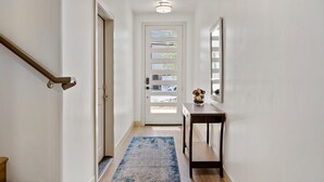 Um corredor com escada de madeira, porta com janelas, uma pequena mesa com objetos decorativos e um tapete azul.