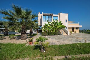 A modern two-story house with a stone patio, palm trees, and a clear blue sky.