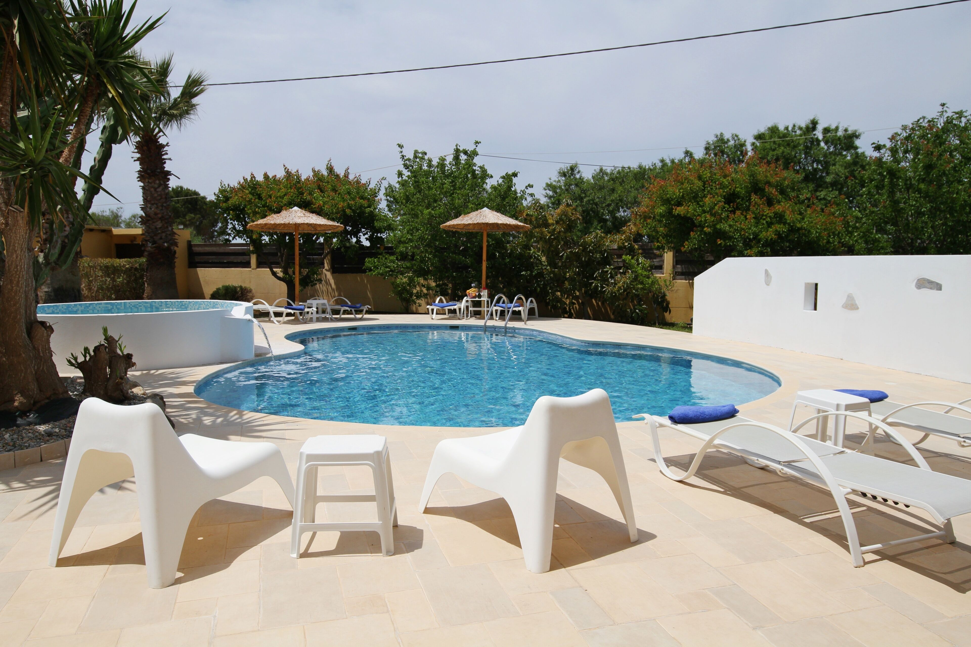 Ein Swimmingpool mit Liegestühlen und einem kleinen Tisch, umgeben von Palmen und einer weißen Mauer.