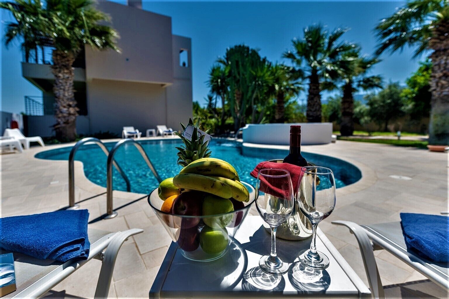 A poolside table with a fruit bowl, wine glasses, and a bottle, overlooking a swimming pool with lounge chairs and palm trees.