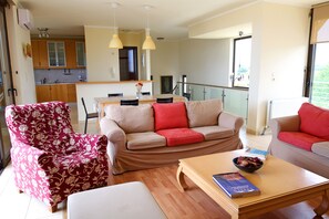 Uma sala de estar moderna com sofá, poltrona e mesa de centro. Uma área de jantar com mesa e cadeiras é visível ao fundo.