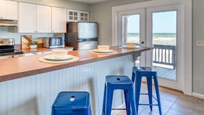 Una cocina con armarios blancos, un refrigerador de acero inoxidable y un amplio ventanal con vista a la playa.