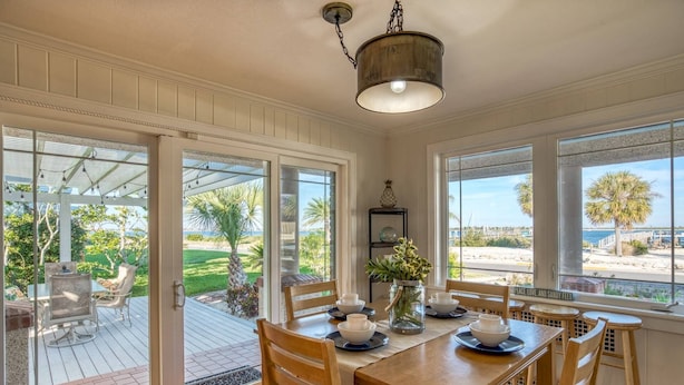A dining area with a wooden table and chairs, a vase with flowers, and a view of the beach and palm trees through large windows. - Waterfront Paradise (St. Augustine)
