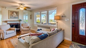 A living room with a fireplace, a ceiling fan, a sofa, a coffee table, and a view of the outdoors through large windows. - Waterfront Paradise (St. Augustine)