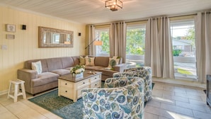 A living room with a beige sofa, a patterned armchair, a coffee table, and a side table. There is a large mirror on the wall and a window with curtains. - Waterfront Paradise (St. Augustine)