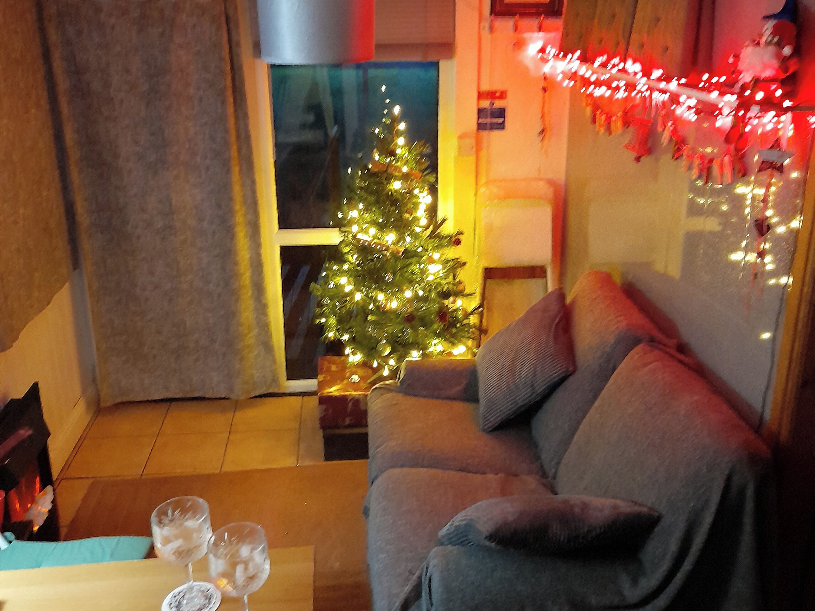 Una sala de estar acogedora con un sofá gris, un pequeño árbol de Navidad y una mesita de vidrio con dos copas de vino.