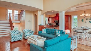 A spacious living room with a large sofa, two patterned ottomans, and a glass coffee table. A kitchen with red cabinets and a dining area with a chandelier are visible in the background. - Tuckers Hideaway at Seagate (St. Augustine)