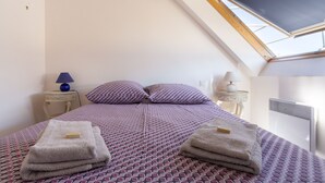 Ein Schlafzimmer mit schrägverdeckter Decke, einem Bett mit gemusterten Bettbezügen, zwei Nachttischlampen und Handtüchern auf dem Bett.