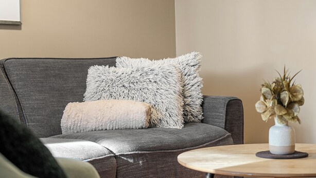 Un canapé gris agrémenté de coussins moelleux blancs et beige, une petite table ronde en bois sur laquelle est posé un vase de fleurs séchées, et un mur beige uni en arrière-plan.