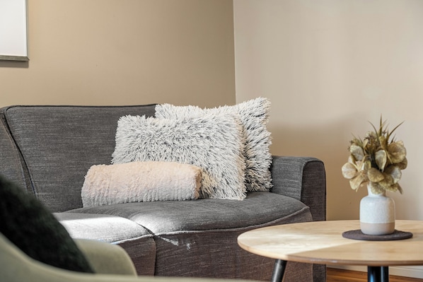 A grey sofa with fluffy white and beige cushions, a small round wooden table with a vase of dried flowers, and a plain beige wall in the background. - Spacious 3 Beds, Stampede, Saddledome, A/C, Underground Parking (Calgary)