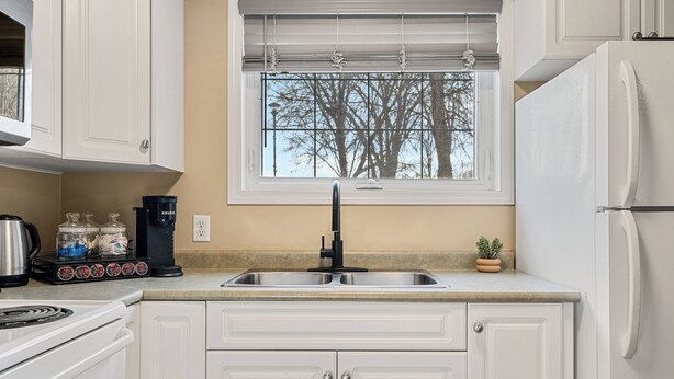A kitchen with white cabinets, a stainless steel sink, a black faucet, a window with blinds, and a refrigerator. - Spacious 3 Beds, Stampede, Saddledome, A/C, Underground Parking (Calgary)