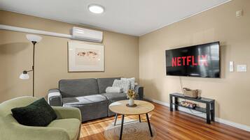 A living room with a grey sofa, a green armchair, a round coffee table, a flat-screen TV, and a wall-mounted air conditioner.