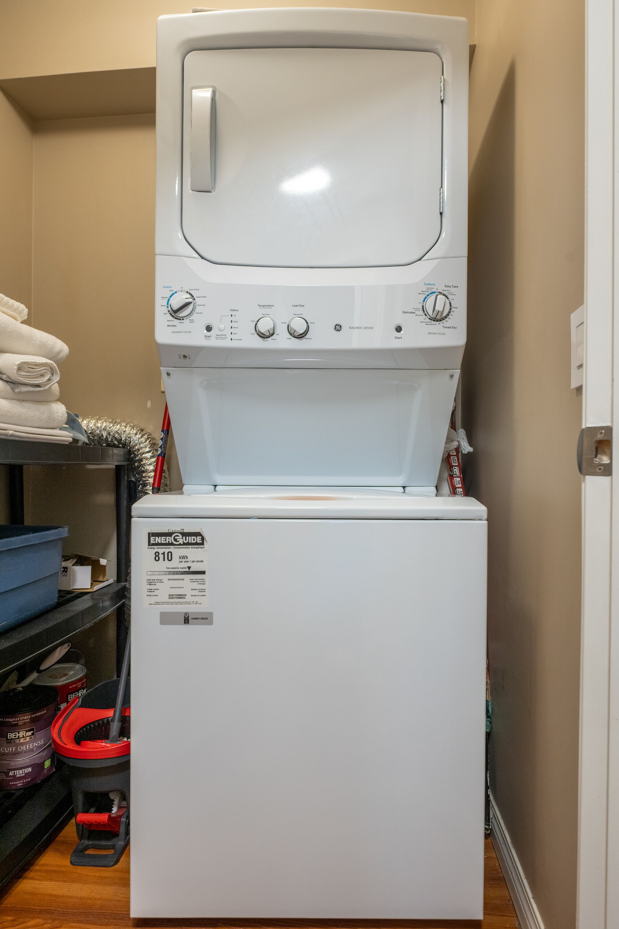 A front-loading washer and dryer unit with a control panel, placed in a laundry room with shelves and cleaning supplies.