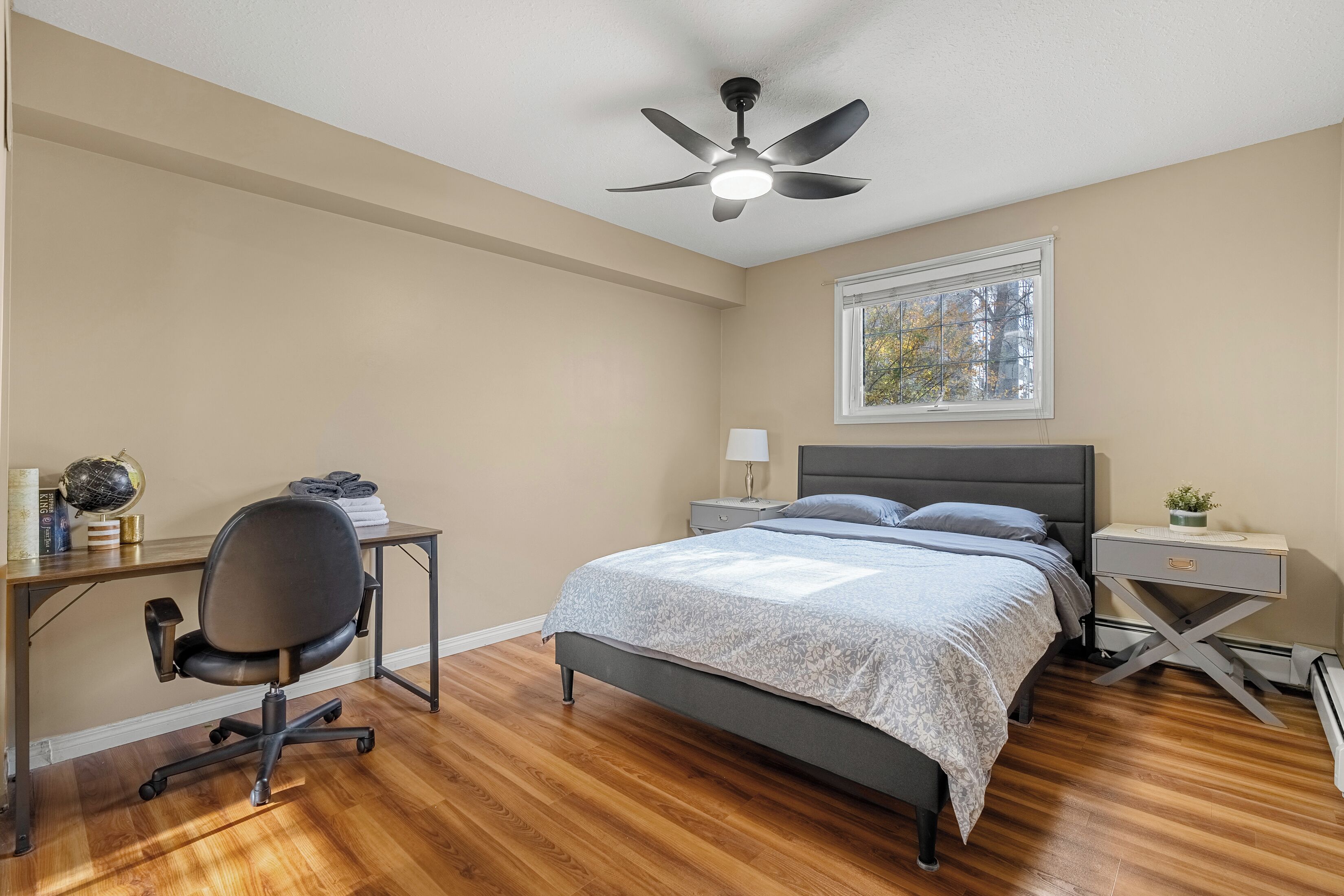 Une chambre avec un lit, un bureau et une chaise, une table de chevet, un ventilateur de plafond et une fenêtre donnant sur des arbres.