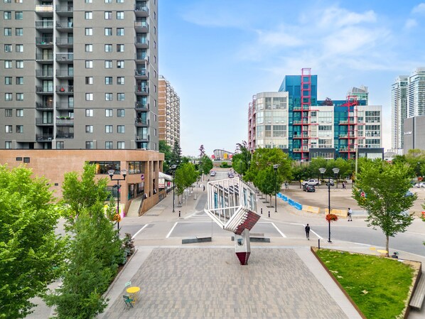 A modern urban square with a prominent sculpture, surrounded by high-rise buildings and a food store.