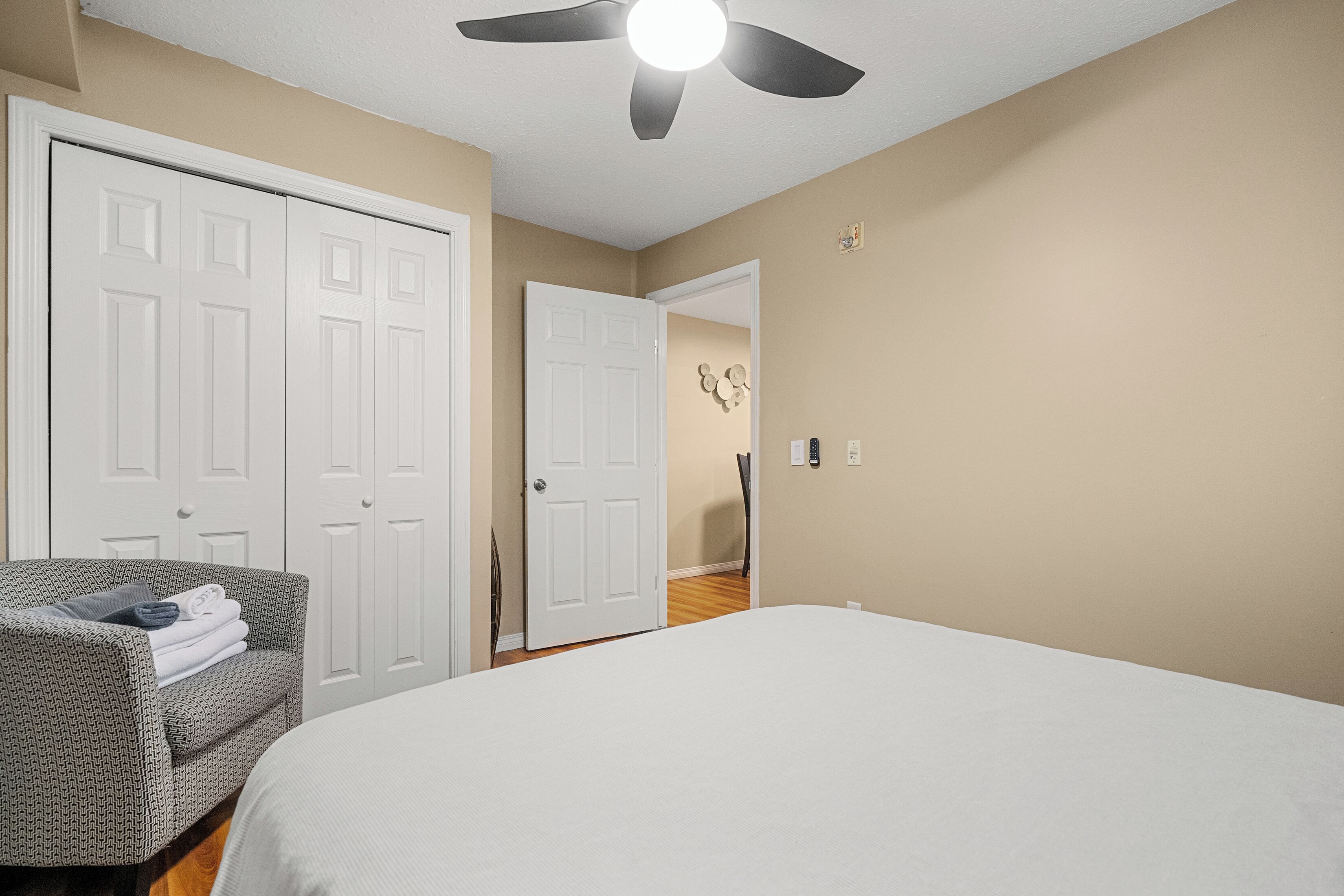 A bedroom with a bed, a chair with folded clothes, a ceiling fan, and white doors.