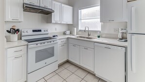 A modern kitchen with white cabinetry, a stainless steel range hood, a dishwasher, and a window with blinds.