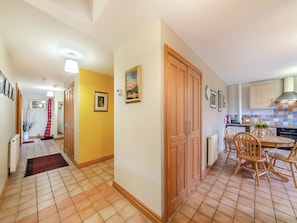 Un corridoio conduce a una cucina con pavimento piastrellato, porte in legno e un'area pranzo con sedie.
