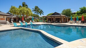 A swimming pool with lounge chairs and umbrellas, surrounded by palm trees and a building with a covered area. - Mauna Lani Villages luxurious Villa with Private Beach Access! (Waimea)