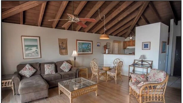 Een ruime woonkamer met een hoge plafond, houten balken, een bank, twee rieten fauteuils, een glazen salontafel, en een eethoek met een tafel en stoelen.