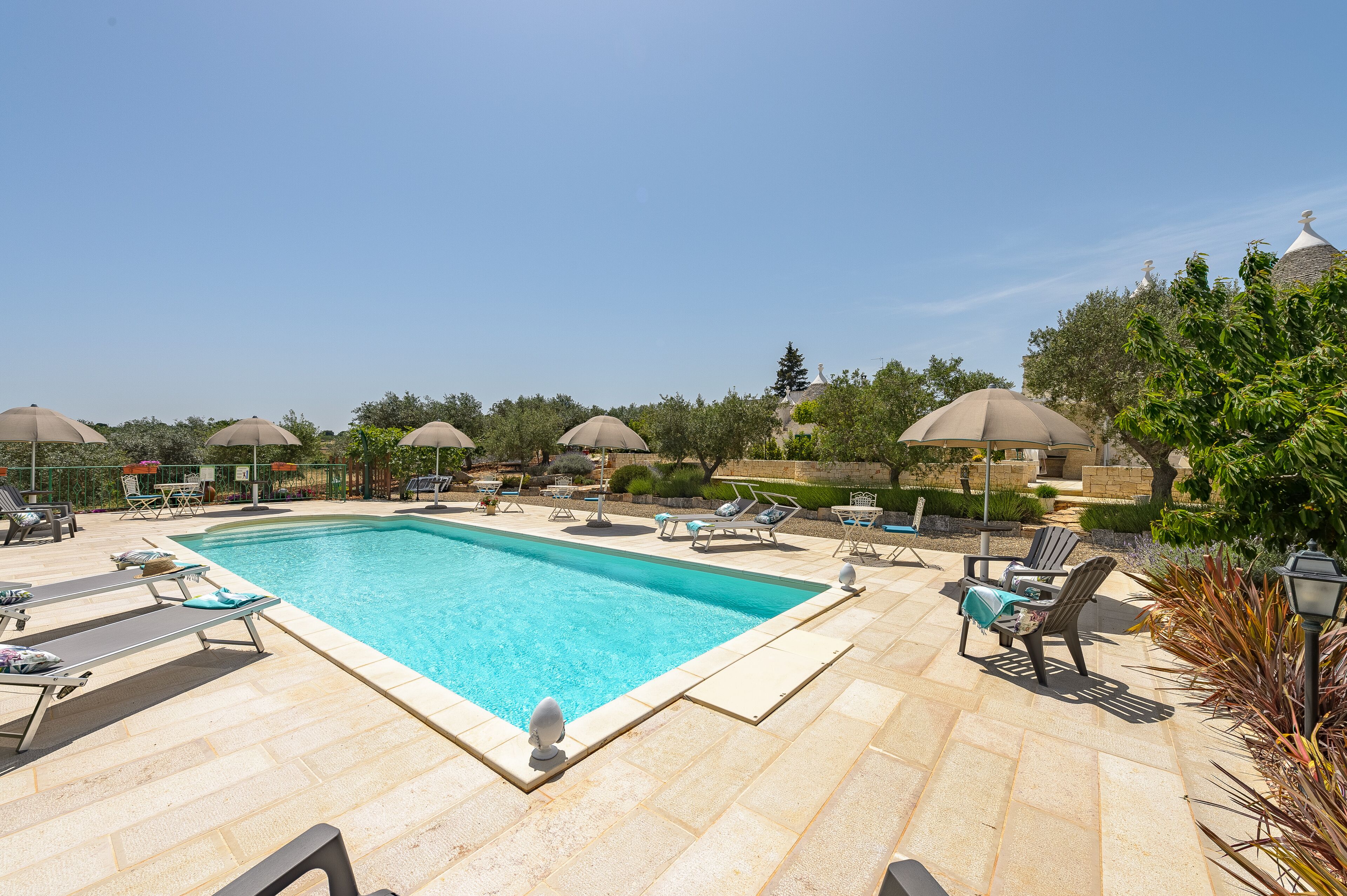 Une piscine rectangulaire, agrémentée de chaises longues et de parasols, entourée d’un jardin aménagé et de dalles de pierre.