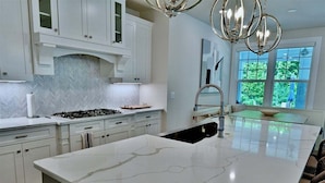A modern kitchen with white cabinetry, a large island, a gas stove, and a view of greenery through the windows.