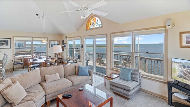 A spacious living area with a large sofa, a coffee table, and a TV on a stand. There is a dining area with a table and chairs, and large windows offering a view of the sea. - Soundfront Dog Friendly w/ Hot Tub, Dock, Private Windsurfing Access, Foosball (Avon)