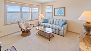 A living room with a view of the sea, a wicker chair, a striped sofa, and a glass coffee table. - Soundfront Dog Friendly w/ Hot Tub, Dock, Private Windsurfing Access, Foosball (Avon)