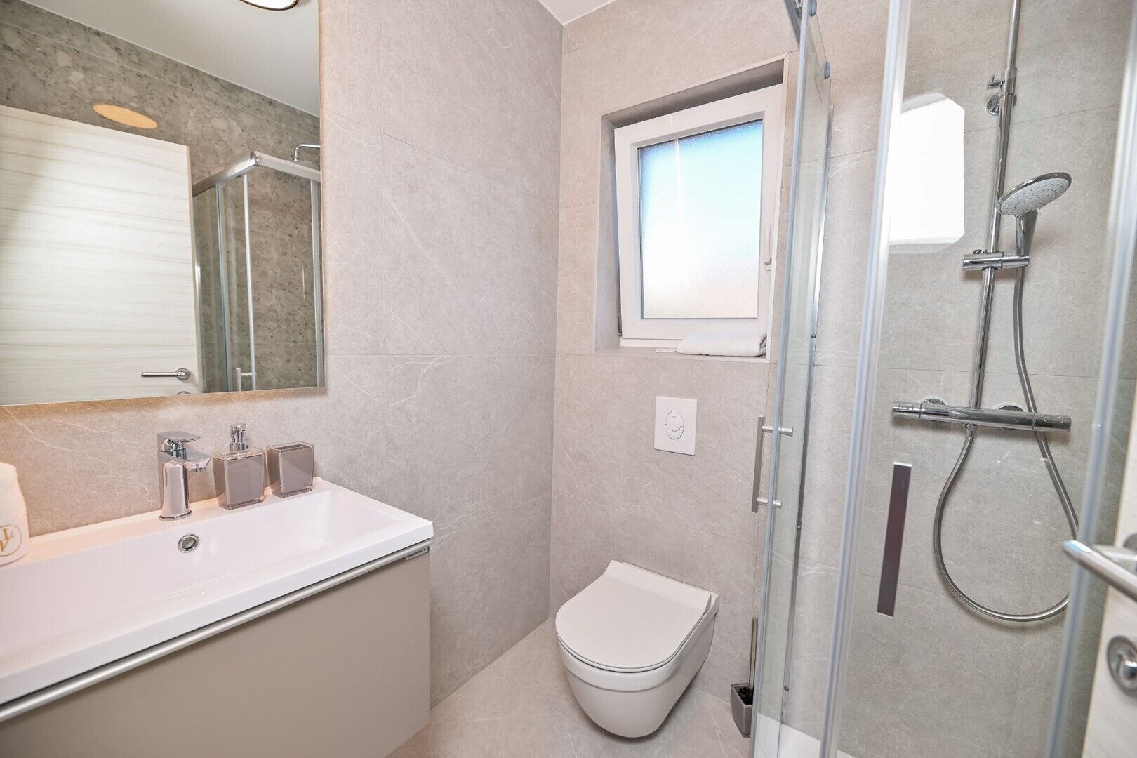 Et moderne badeværelse med et brusebad bag glas, et hvidt toilet og en vaskebord med et spejl og to håndvaske.