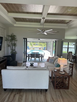 Uma sala de estar ampla com ventilador de teto, um sofá grande, uma mesa de centro e vista para uma área de piscina externa.