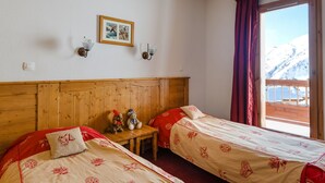 A room with two beds, a wooden headboard, a small table with two teddy bears, a framed picture on the wall, and a window with a view of snow-covered mountains.