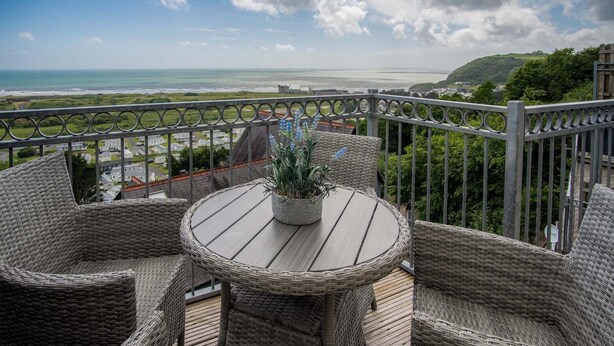A balcony with wicker furniture, a potted plant, and a view of the sea and greenery.