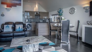 A modern living room with a marble coffee table, a sofa with decorative pillows, a dining area with a table and chairs, and a kitchen with a tiled backsplash.