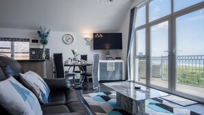 A modern living room with a black leather sofa, a dining area with a round table, and a balcony with a view.