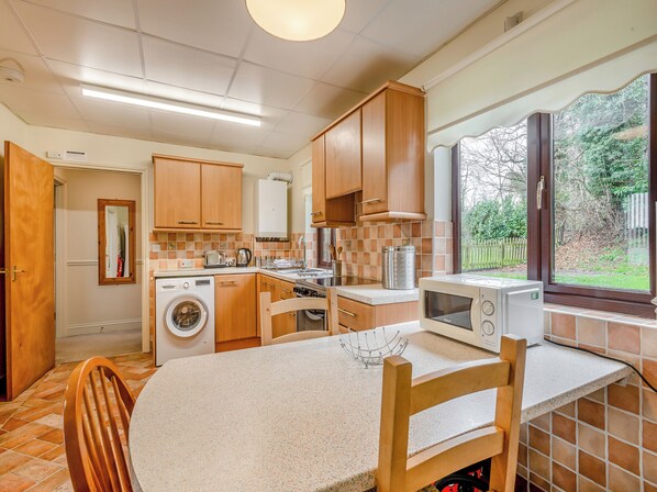A compact kitchen with wooden cabinets, a washing machine, a microwave, and a dining area with a table and chairs.