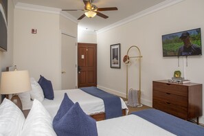 Une chambre d’hôtel avec deux lits, un ventilateur de plafond, une télévision, une commode en bois et une porte donnant sur une autre pièce.