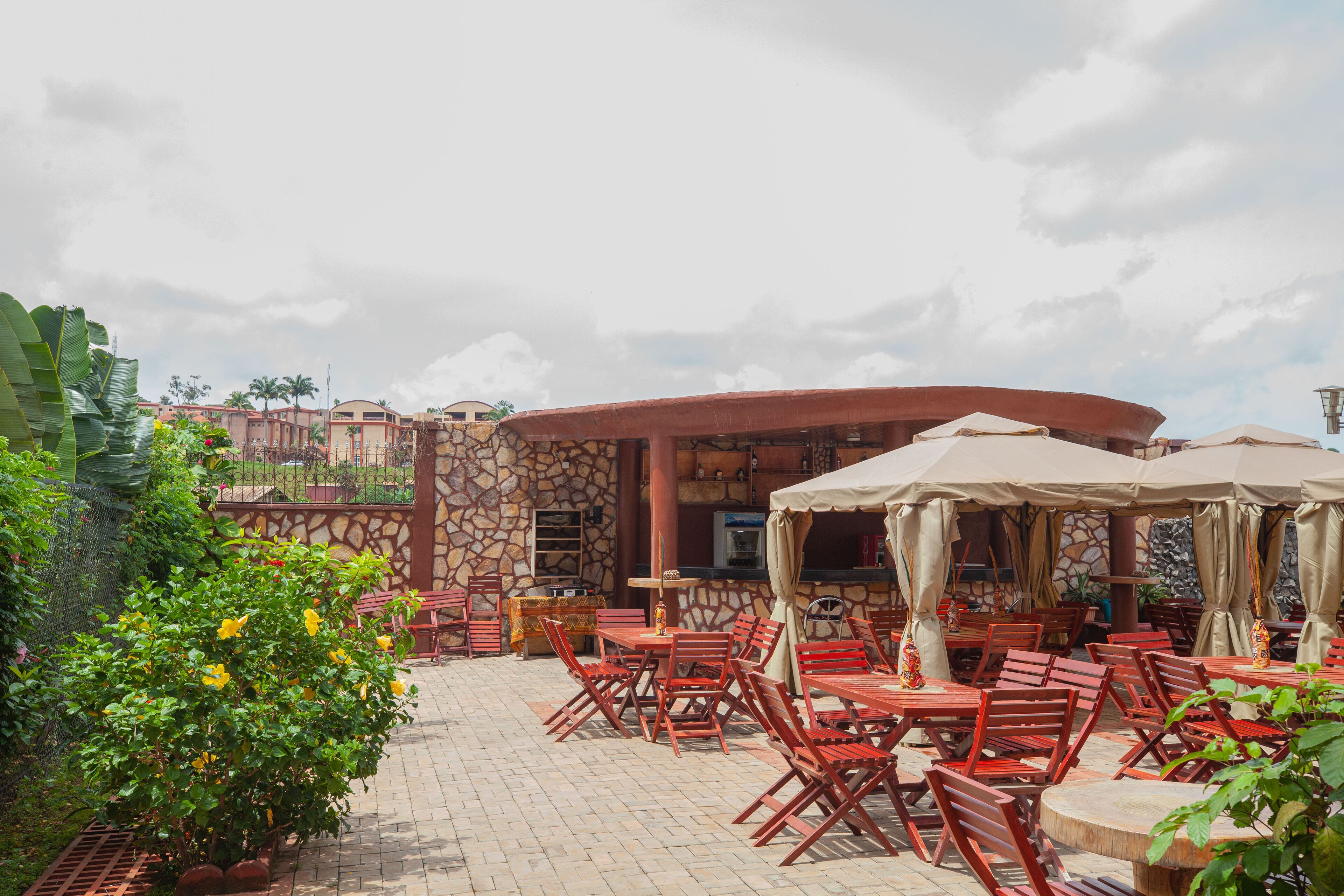 An outdoor patio area with red wooden tables and chairs, beige umbrellas, and a stone wall backdrop.