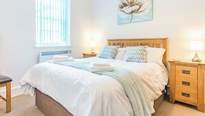 A neatly made bed with white linens and a light blue throw blanket, a wooden nightstand with a lamp, and a window with blinds. - Claro Cottage - UK37450 (Pateley Bridge, near Harrogate)