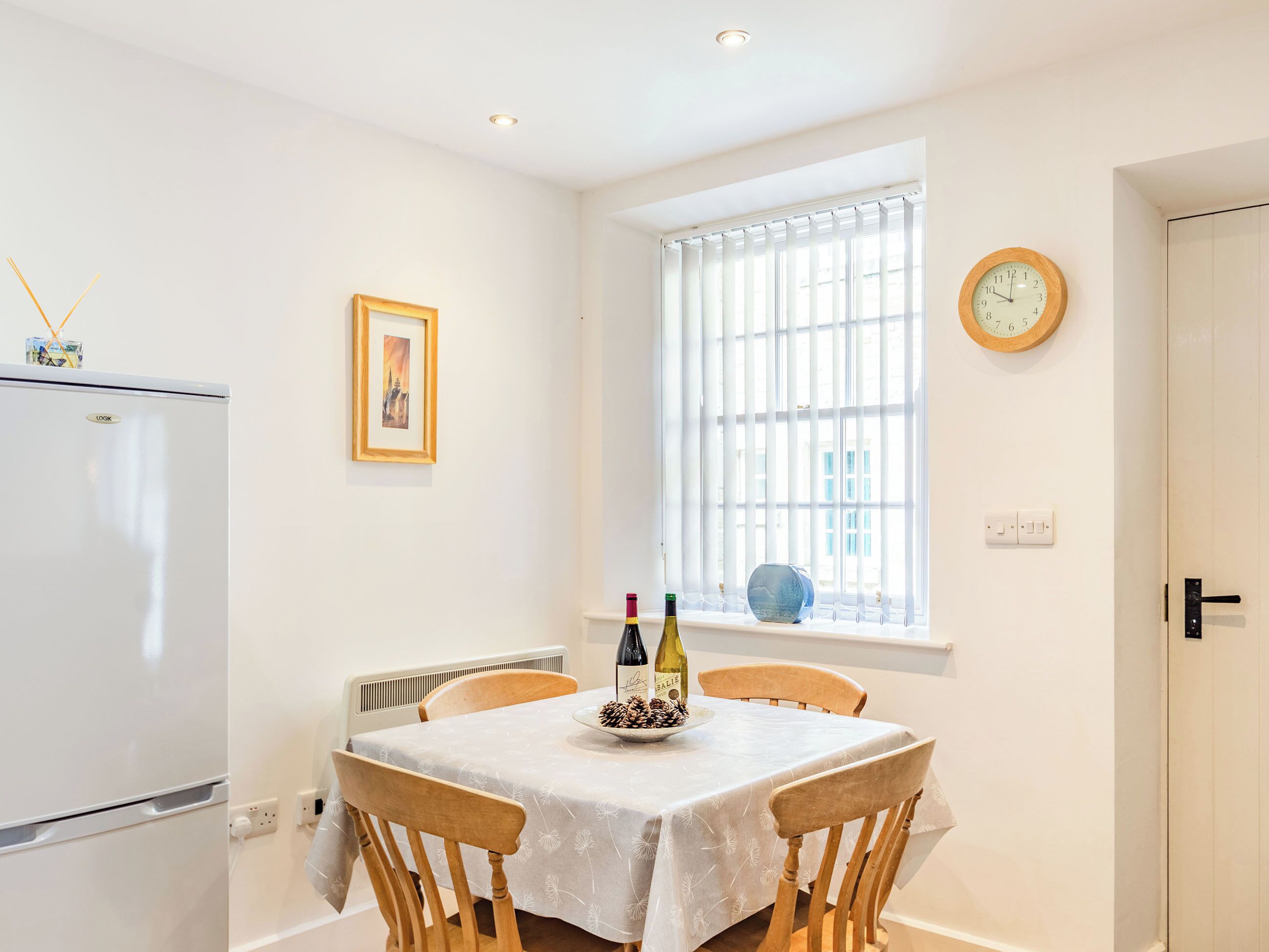 Un comedor con una mesa dispuesta para dos, una ventana con persianas, un reloj de pared y un refrigerador.