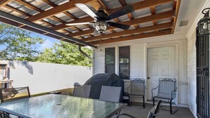 Une terrasse extérieure avec une table en verre, des chaises en métal, un ventilateur de plafond et un barbecue.