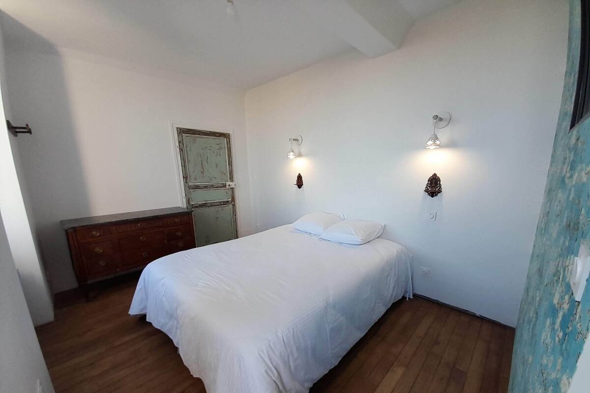Quarto com piso de madeira, cama com roupa de cama branca, cômoda e duas luminárias fixadas na parede.