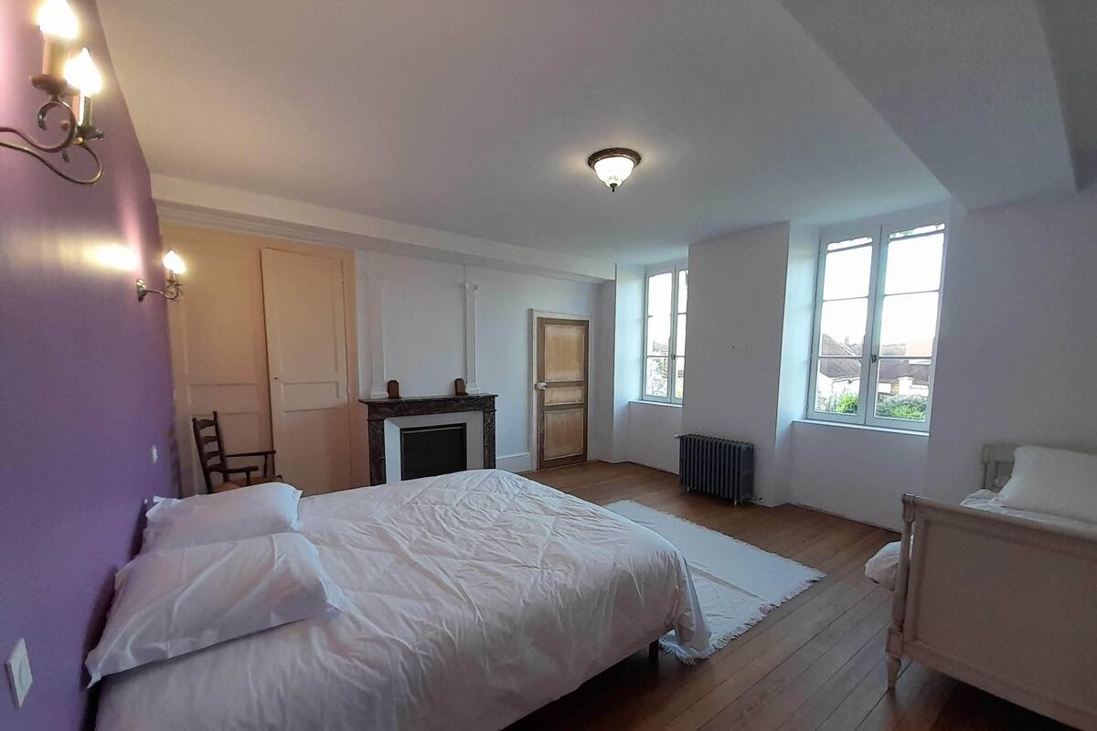 Um quarto com cama, uma cadeira, lareira e janelas amplas.