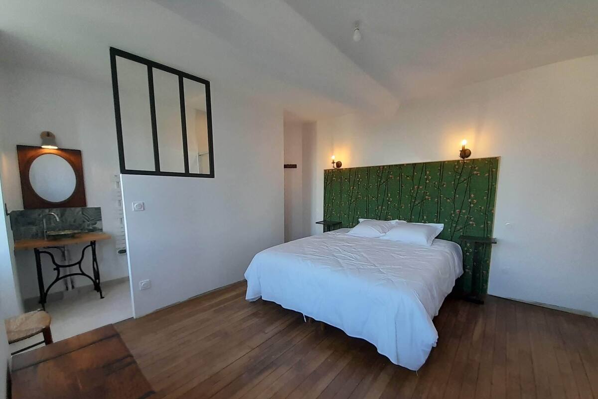 Quarto com cama, mesa de cabeceira, espelho fixado na parede e pequena mesa com cadeira.